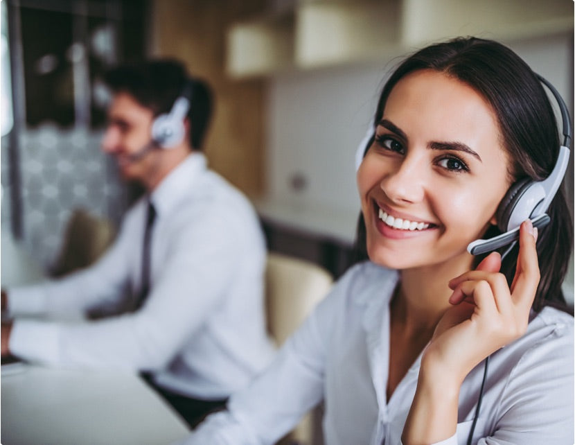 Zwei Kundenservice-Mitarbeitende mit Headsets sitzen an Schreibtischen; im Vordergrund eine lächelnde Frau in weißer Bluse.