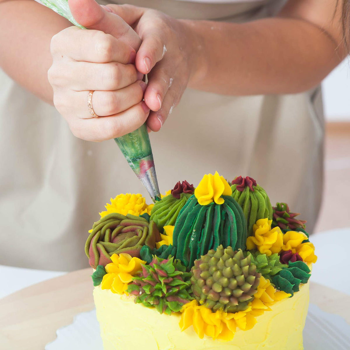 Spritzbeutel-Set mit Tüllen - 67-tlg. zeigt Hand, die mit grünem Spritzbeutel bunte Kakteen und Blumen auf Torte verziert.