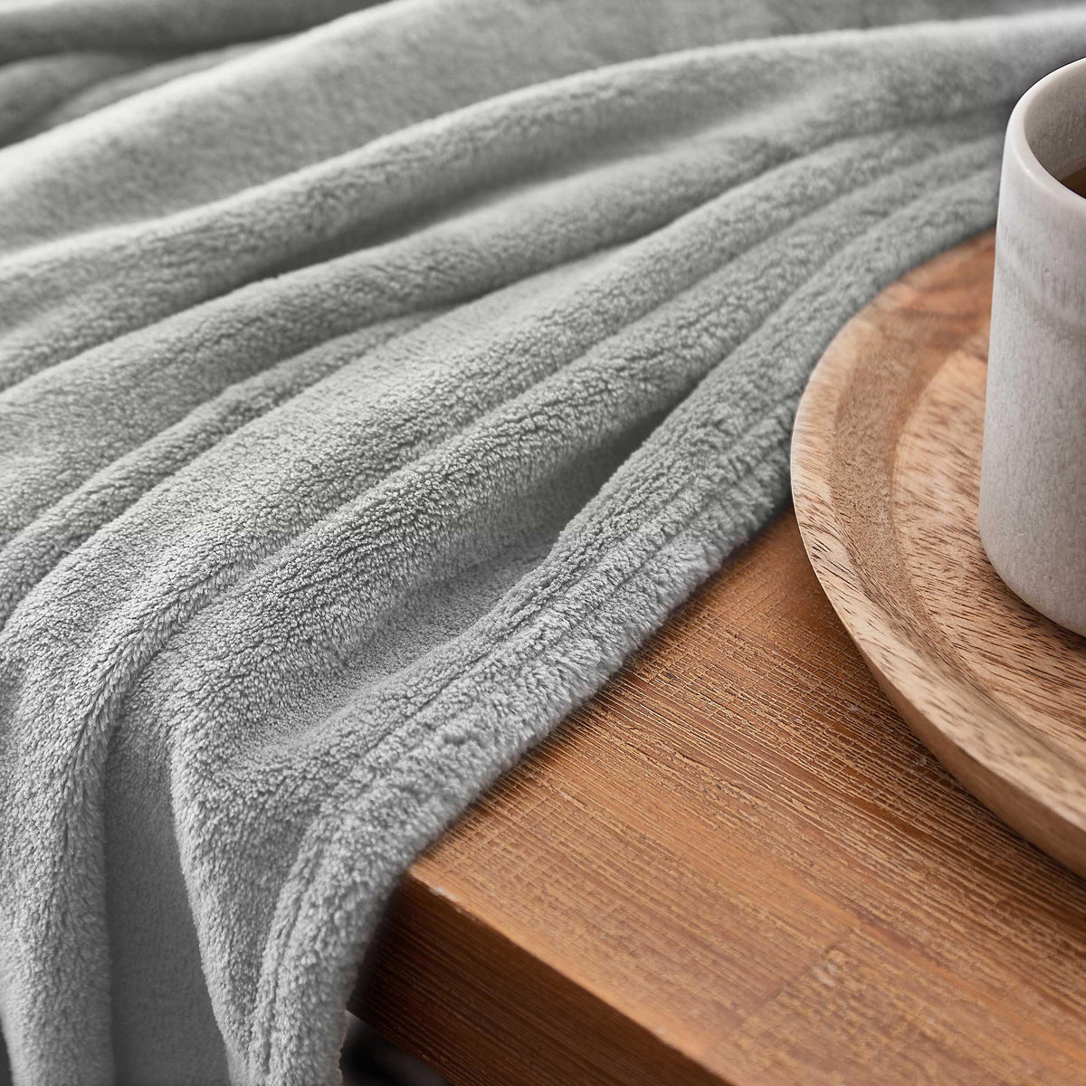 Kuscheldecke - 220x240 cm - Sofadecke & Tagesdecke - Hellgrau aus weichem Coral-Fleece auf Holztisch mit Tasse.