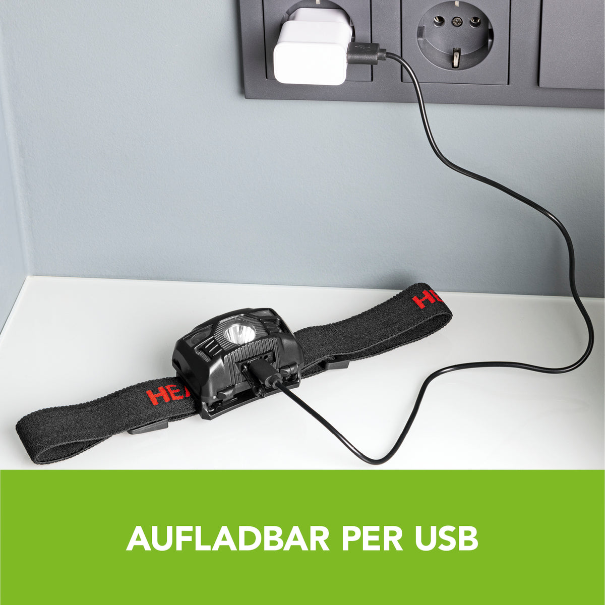 Stirnlampe schwarz/rot mit USB-Ladekabel auf weißem Tisch vor Steckdose, wiederaufladbar und mit verstellbarem Kopfband