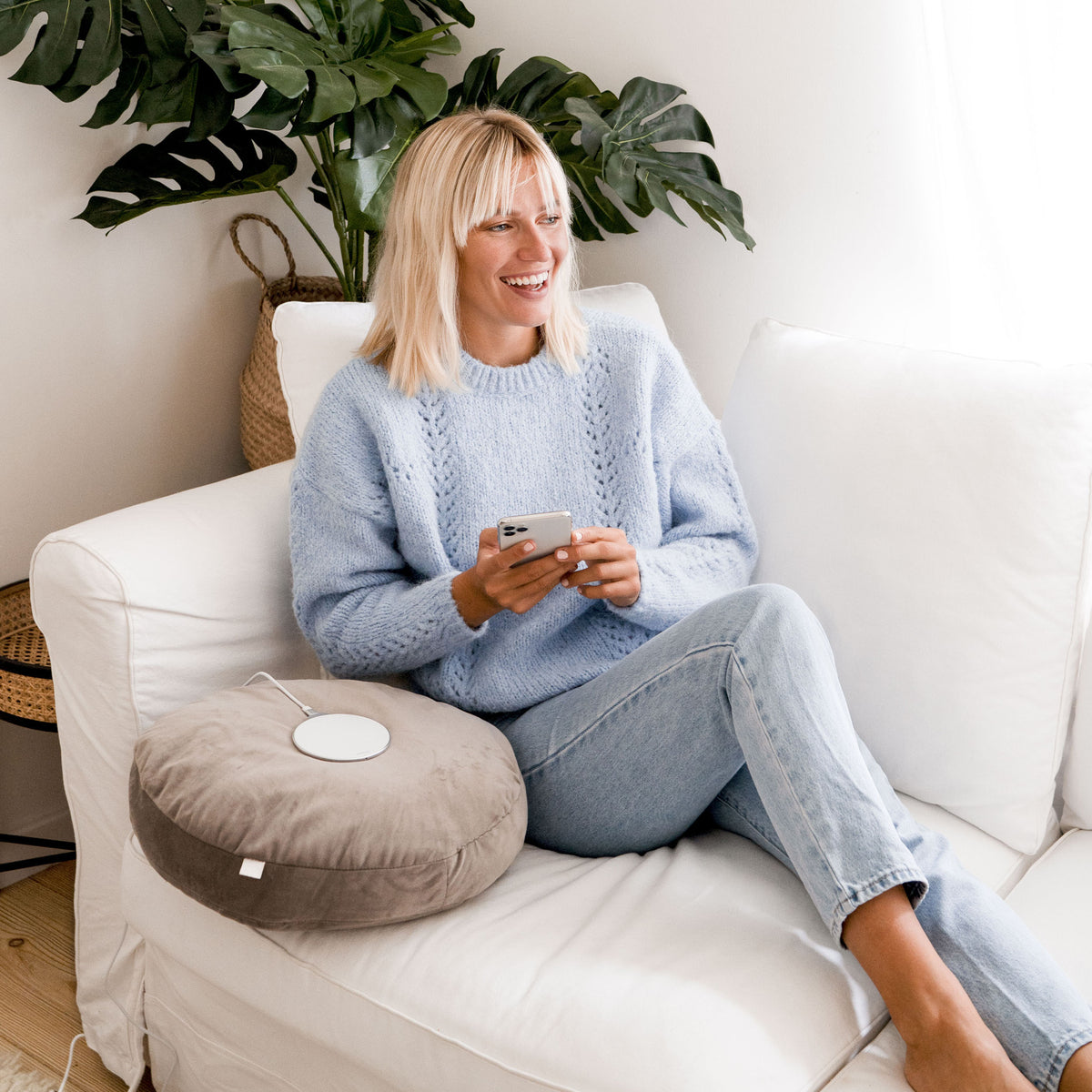 Frau sitzt entspannt auf Sofa, nutzt Wireless Ladegerät Aura lite 15W schwarz zum kabellosen Laden ihres Smartphones.
