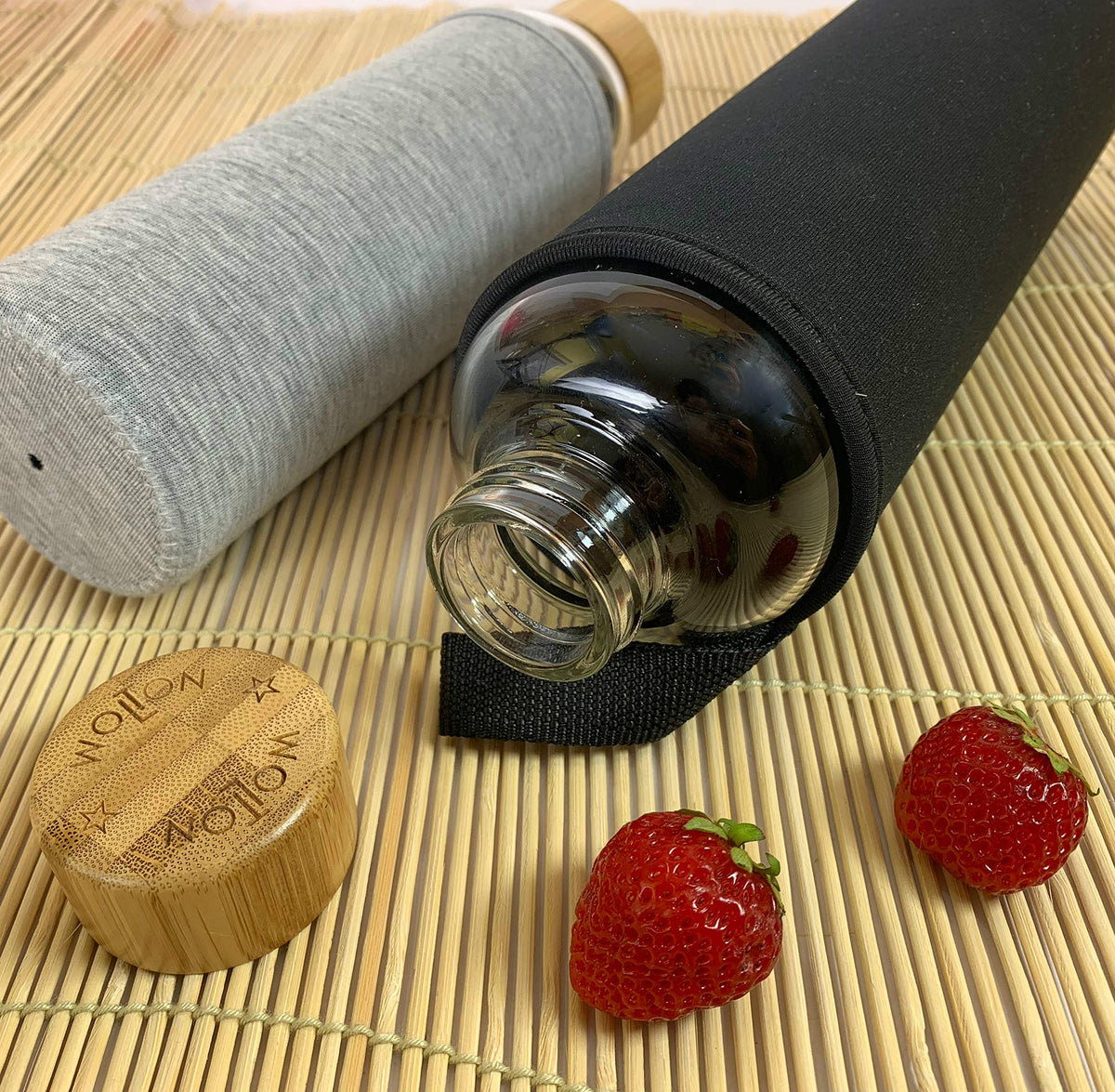 Trinkflasche mit Bambusdeckel - 550 ml - Grau, mit grauem Stoffüberzug, geöffnet und zwei frischen Erdbeeren auf Bambusmatte.