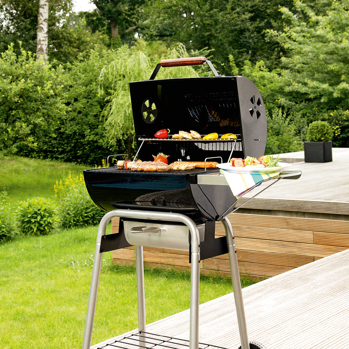 Holzkohle-Barrelgrill mit lackiertem Deckel, emailliertem Rost, Warmhalterost und Temperaturanzeige im Garten auf Terrasse.