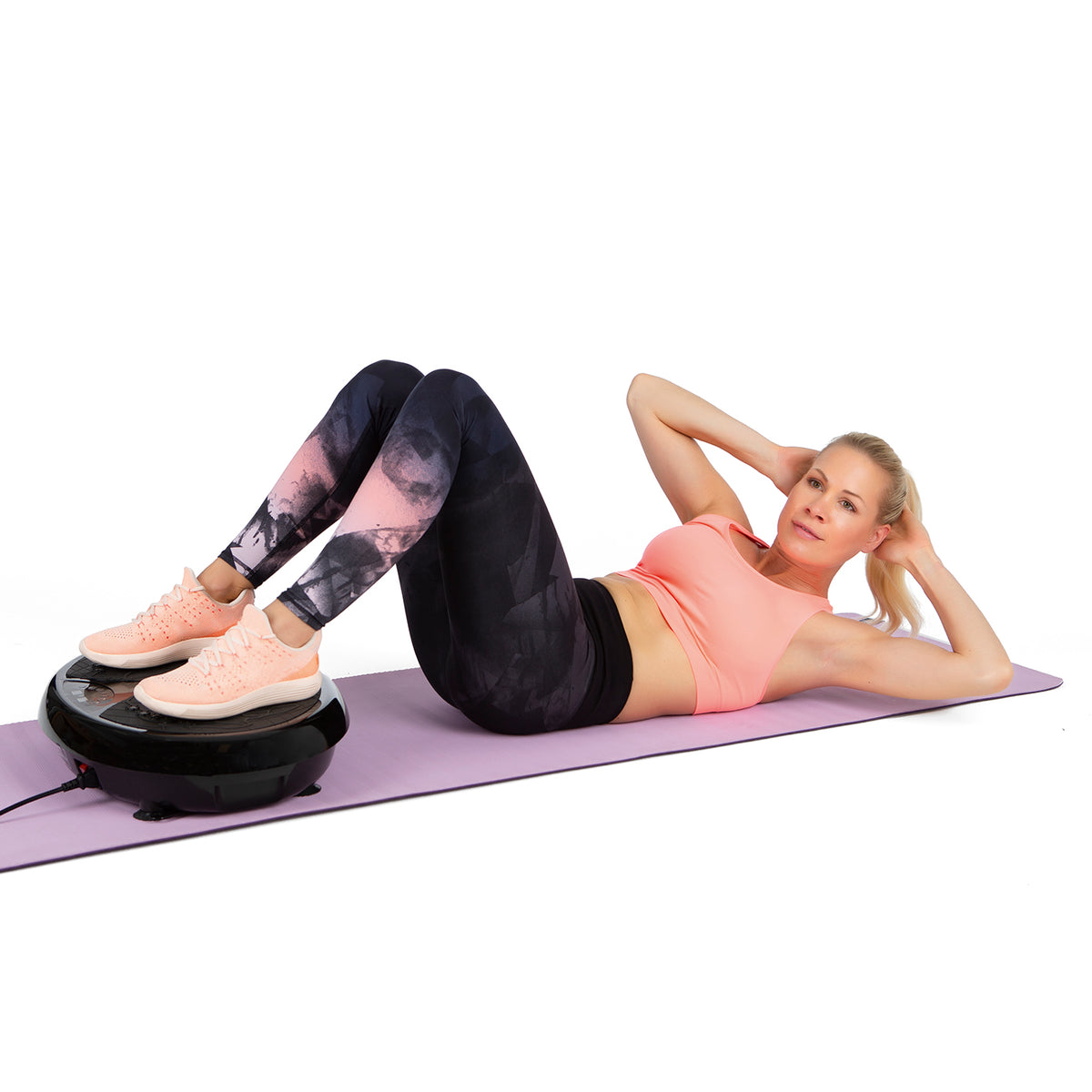 Frau trainiert auf Vibrationstrainer auf Yoga-Matte mit angewinkelten Beinen und Händen hinter dem Kopf in Sportkleidung.