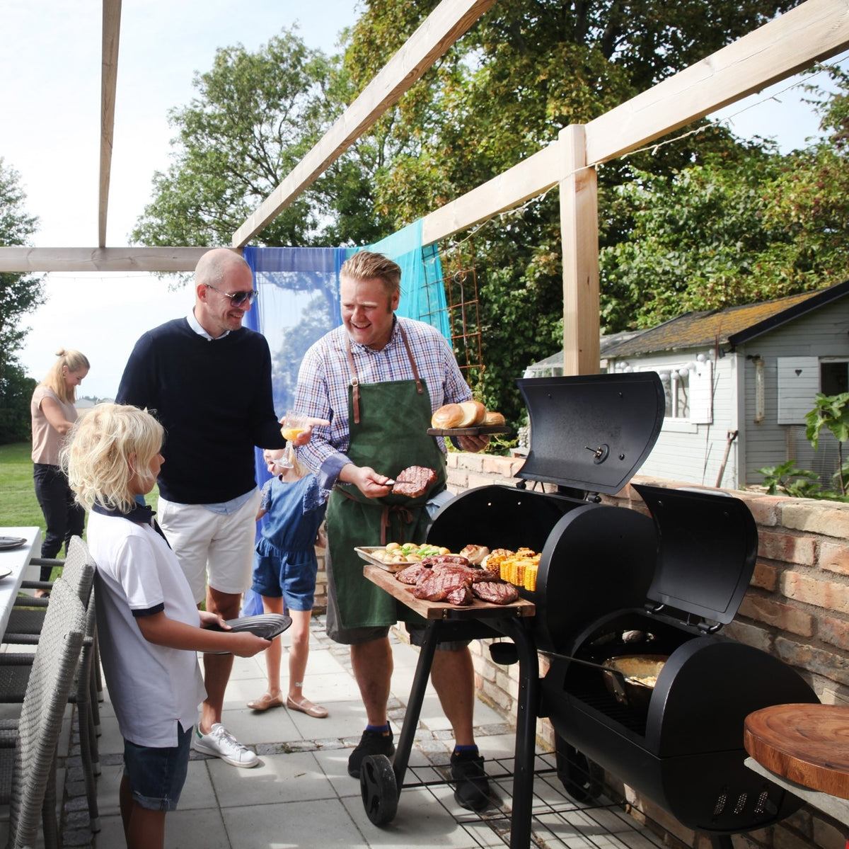 Smoker Vinson 200 mit 4-in-1 Funktion, abgerundetem Design und Edelstahlgriffen beim Grillen im Garten mit Familie.